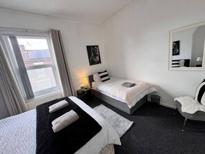 Habitación de hotel con dos camas, una ventana con cortinas, una mesita de noche con lámpara, un cuadro en la pared y un espejo.