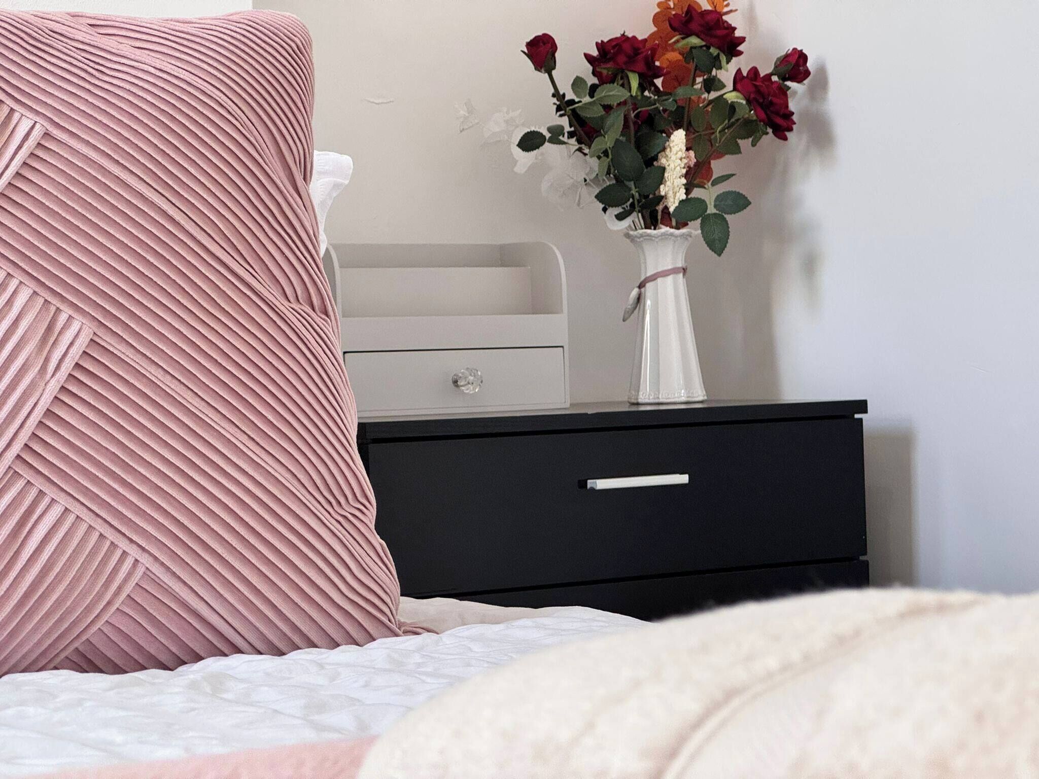 Een bed met een roze kussen, een witte vaas met bloemen en een nachtkastje met lades.
