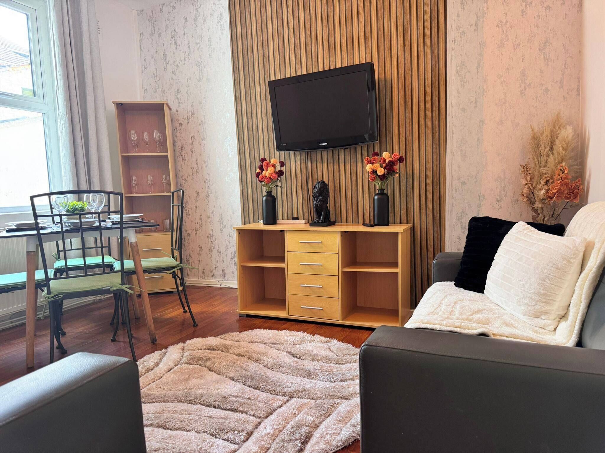 A living room with a flat-screen TV, a sofa, a coffee table, a dining area with chairs and a table, and a bookshelf.