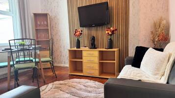 A living room with a flat-screen TV, a sofa, a coffee table, a dining area with chairs and a table, and a bookshelf.