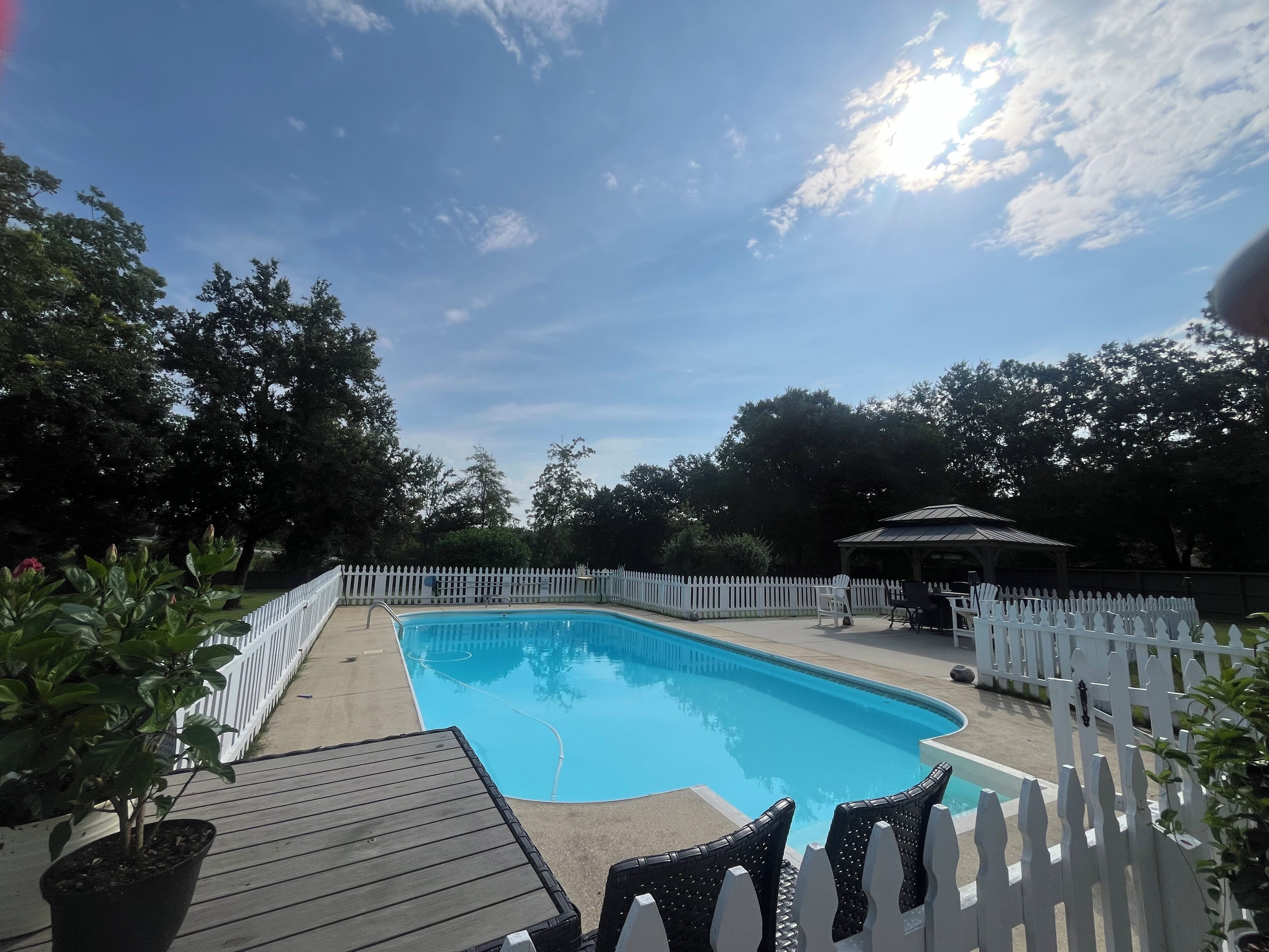 A rectangular swimming pool surrounded by a white picket fence, with a covered seating area and potted plants nearby.