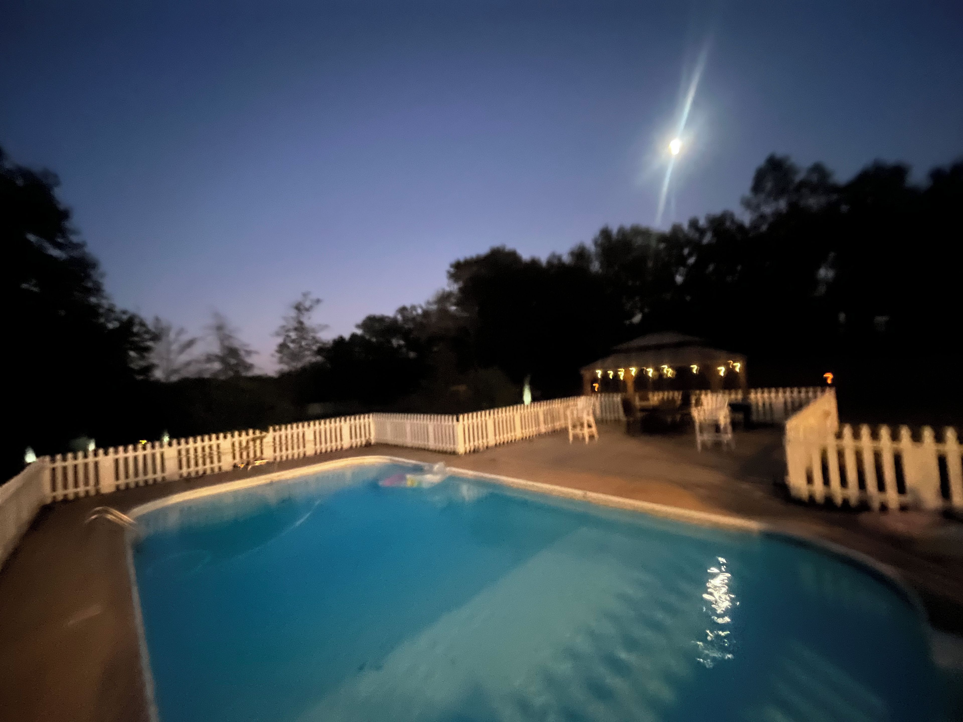 A swimming pool with a white fence, a gazebo, and trees in the background.