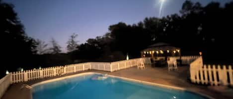 A swimming pool with a white fence, a gazebo, and trees in the background.
