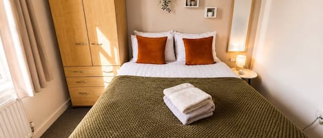 A hotel room with a bed, a wardrobe, a bedside table with a lamp, and a wall-mounted mirror.
