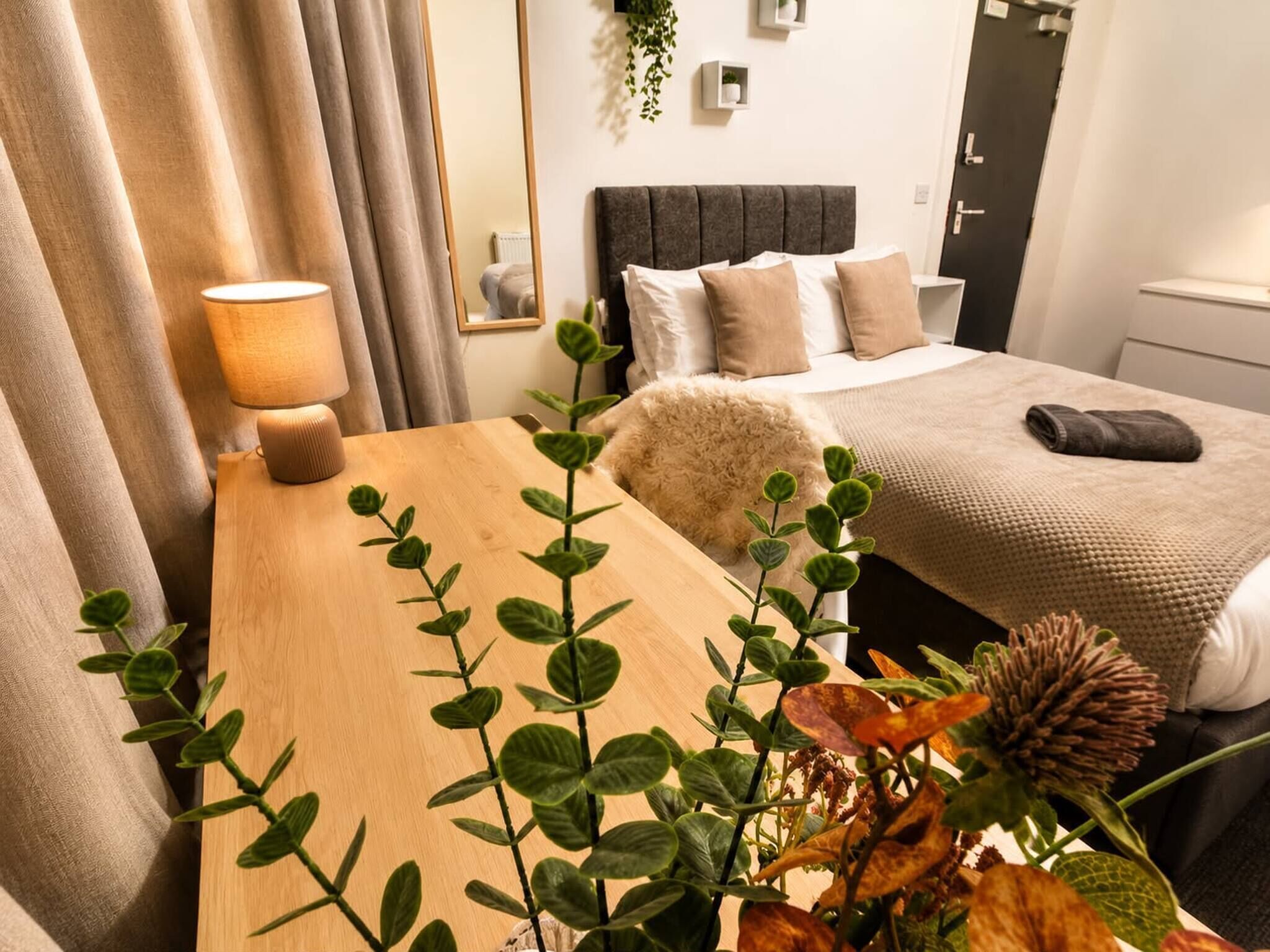 A hotel room with a wooden desk, a lamp, a bed with a grey headboard, and a plant in the foreground.