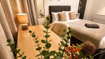 Ein Hotelzimmer mit einem Holztisch, einer Lampe, einem Bett mit grauem Kopfteil und einer Pflanze im Vordergrund.