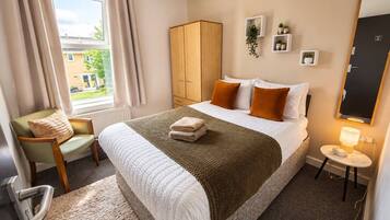 A hotel room with a bed, a chair, a wardrobe, a bedside table with a lamp, and a view of the outside.
