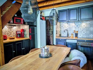 A kitchen with a wooden table, a coffee maker, a refrigerator, and patterned backsplash tiles. - Le Cerf Blanc, appartement de charme proche des pistes, Les Angles (Les Angles)