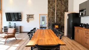 A spacious room with a long wooden dining table, black chairs, a stone fireplace, a kitchen area with a black refrigerator, and a flat-screen TV mounted on the wall. - Casa para 8 Personas a solo 5 km de Corralco (Malalcahuello)