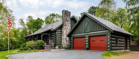 A log cabin with a stone chimney, two red garage doors, and a flagpole with an American flag.