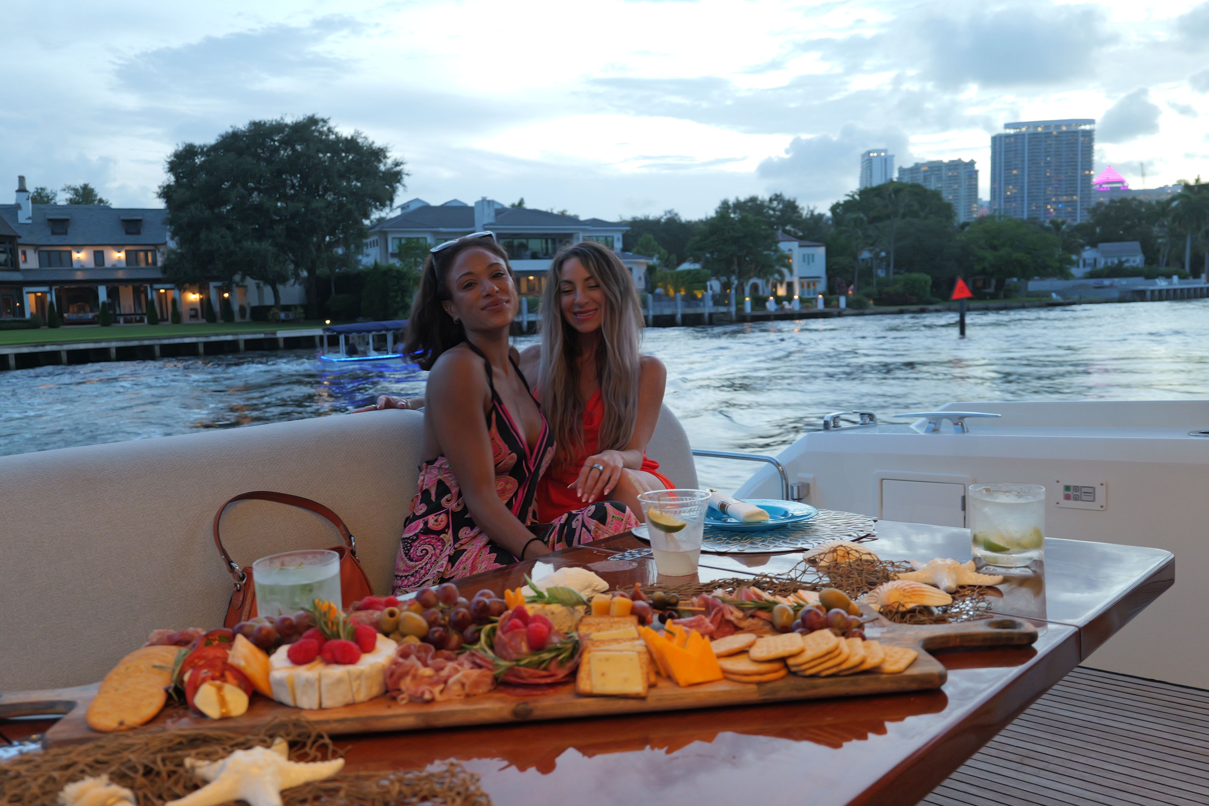 Dos personas en un bote con una mesa de comida y bebidas, un río y edificios al fondo.