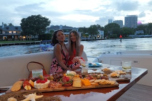 Two people on a boat with a table of food and drinks, a river, and buildings in the background.