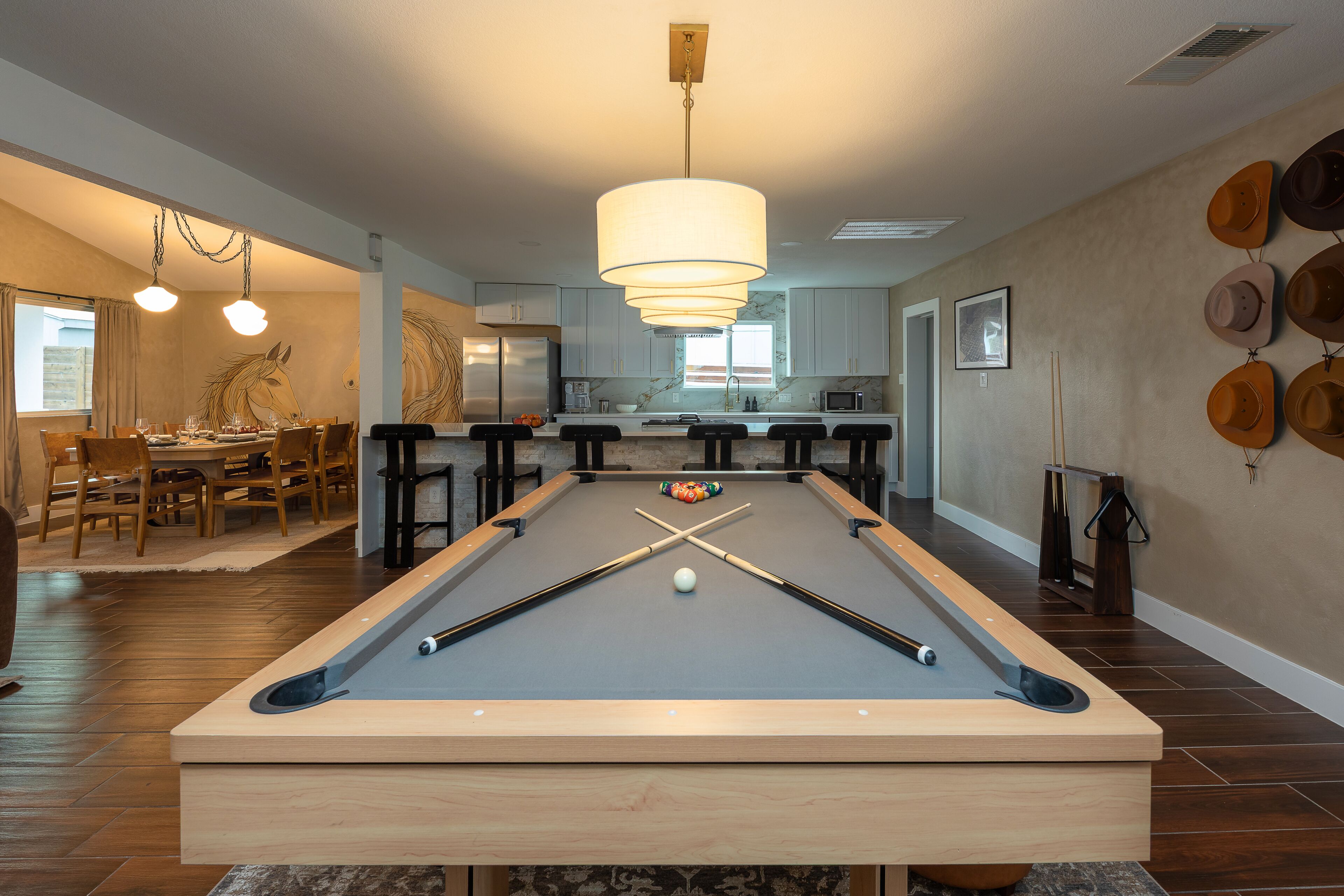 A pool table with a billiard cue, balls, and a pool table in a room with wooden flooring, a dining area, and a kitchen in the background.
