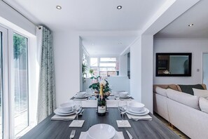 A modern dining area with a table set for six, a bottle of champagne, and a sofa in the background. - *NEW* 3BR Home | Driveway | Garden | Pool Table (West Midlands)