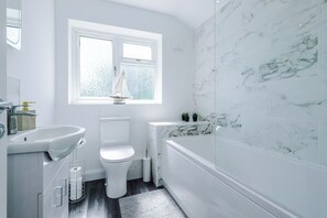 A modern bathroom with a white sink, toilet, and bathtub, marble walls, and a window with a sailboat view. - *NEW* 3BR Home | Driveway | Garden | Pool Table (West Midlands)