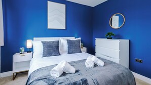 Ein Schlafzimmer mit Bett, weißem Kopfteil, blauen Wänden, einem Nachttisch mit Lampe, einem Schminktisch und einem Spiegel.