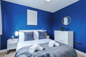 A bedroom with a bed, white headboard, blue walls, a bedside table with a lamp, a dresser, and a mirror. - NEW Relaxing 4BR House with Free WIFI & Parking (Bramhall)