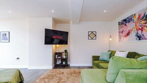 Ein modernes Wohnzimmer mit einem grünen Sofa, einem Flachbildfernseher, der 'NETFLIX' zeigt, einem farbenfrohen abstrakten Gemälde und einer kleinen Pflanze auf einem Holzregal.