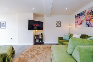 A modern living room with a green sofa, a flat-screen TV displaying 'NETFLIX', a colorful abstract painting, and a small plant on a wooden shelf. - NEW Relaxing 4BR House with Free WIFI & Parking (Bramhall)