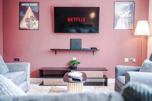Una sala de estar con un televisor de pantalla plana montado en la pared, una mesita con un control remoto y dos sillones.