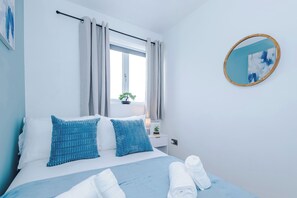 Ein Schlafzimmer mit einem Bett, Kissen, einem Fenster mit Vorhängen, einem Nachttisch mit Lampe, einem Spiegel und einer Wand mit einem Gemälde.
