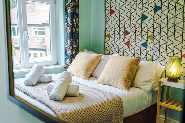 Ein ordentlich bezogenes Bett mit weißen Leinen und mehreren Kissen, ein Fenster mit Vorhängen und einer Wand mit Muster.