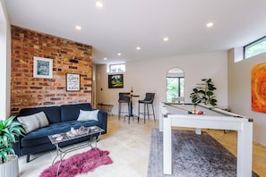 A modern living room with a brick wall, a blue sofa, a pool table, and a dining area with a window. - *NEW* Funky 4BR Home | Sleeps 8 | Driveway | PS4 (Greater Manchester)