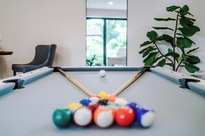 Een pooltafel met ballen, een stoel en een plant in een kamer met een raam.