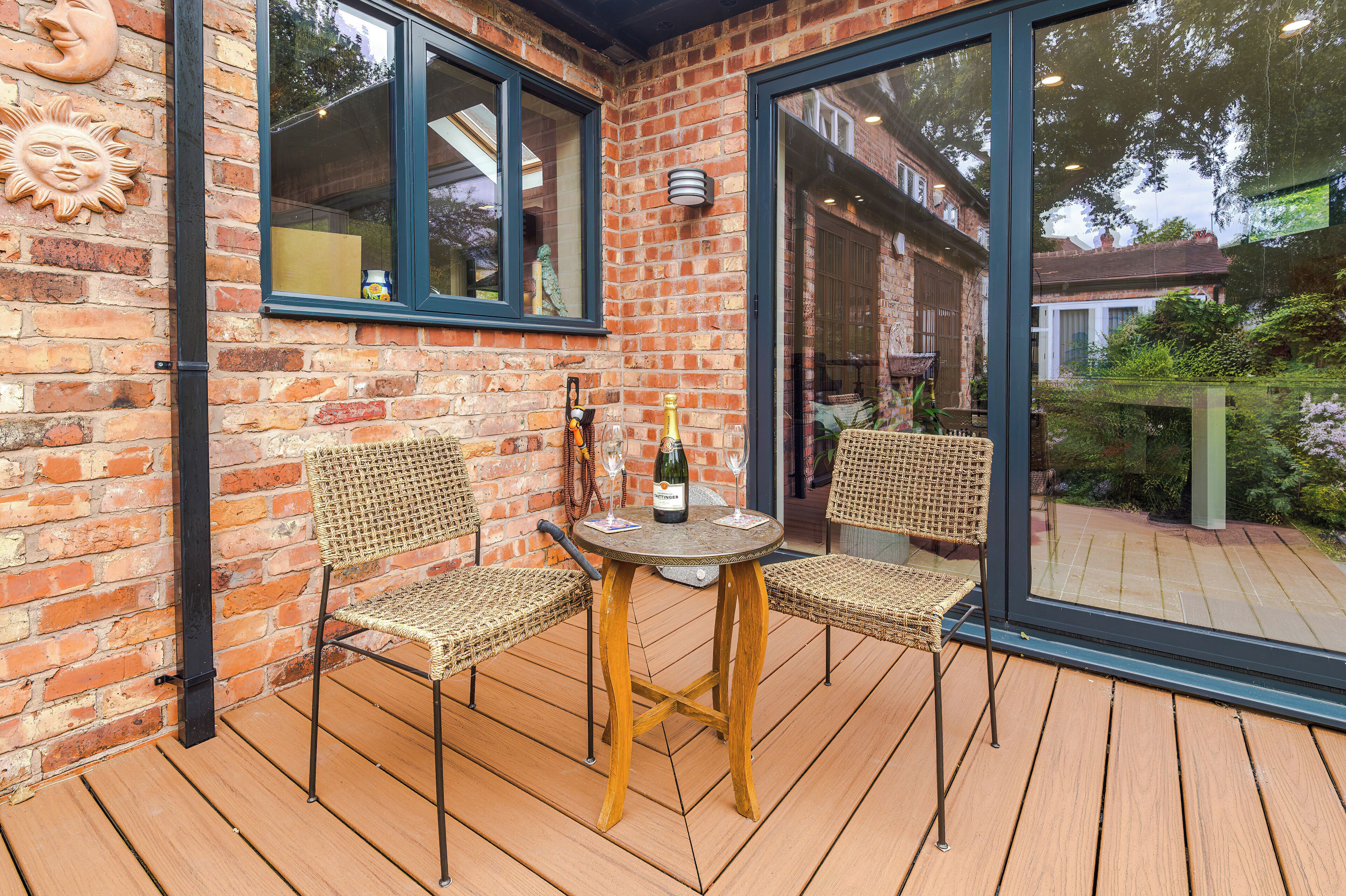A wooden deck with two wicker chairs and a table, a brick wall, and a glass door leading to a garden.