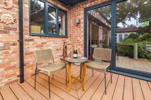 Eine Holzterrasse mit zwei Korbstühlen und einem Tisch, eine Backsteinmauer und eine Glastür, die in einen Garten führt.