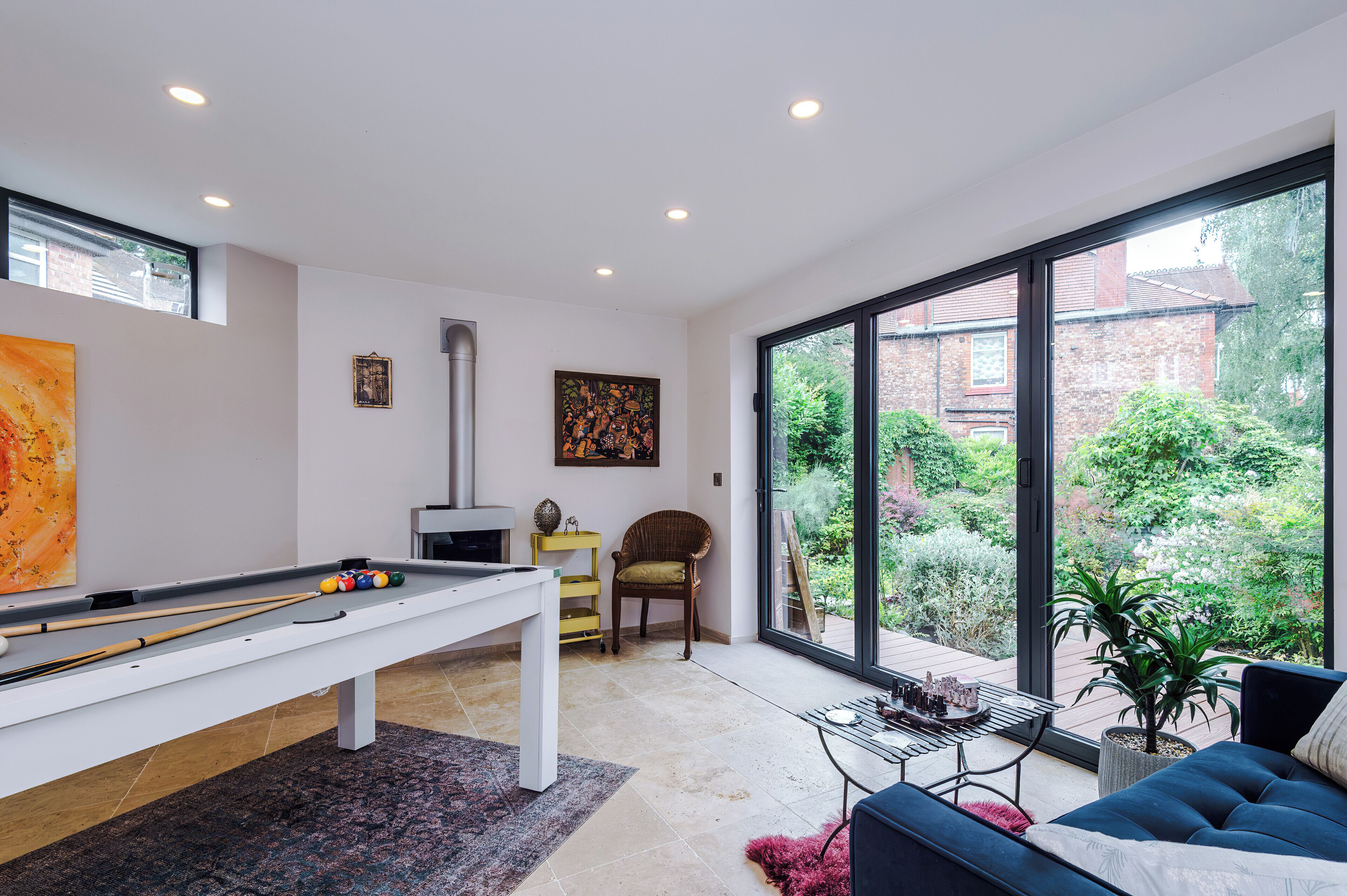 A modern living room with a pool table, a sofa, and a large window offering a view of a garden and a brick building.