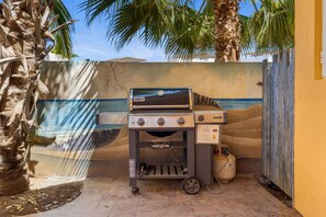 Una parrilla Weber con tanque de gas, un mural azul y blanco, y una palmera.