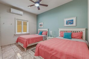 A bedroom with two beds, a ceiling fan, an air conditioner, a window, and two framed pictures on the wall. - Relaxing Family 2BR Condo / Walk to Beach & Pool (El Pescadero)