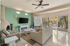 Uma sala de estar com um sofá, uma mesa de centro, uma TV de tela plana, um ventilador de teto e uma porta de vidro deslizante que dá para uma área externa com um jardim de cactos.