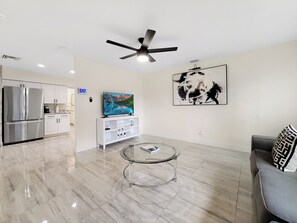 A modern living room with a glass coffee table, a gray sofa, a ceiling fan, a kitchen area with stainless steel appliances, and a large abstract painting on the wall. - Centric Vacation Home with Tiny House near Beaches (Tampa)