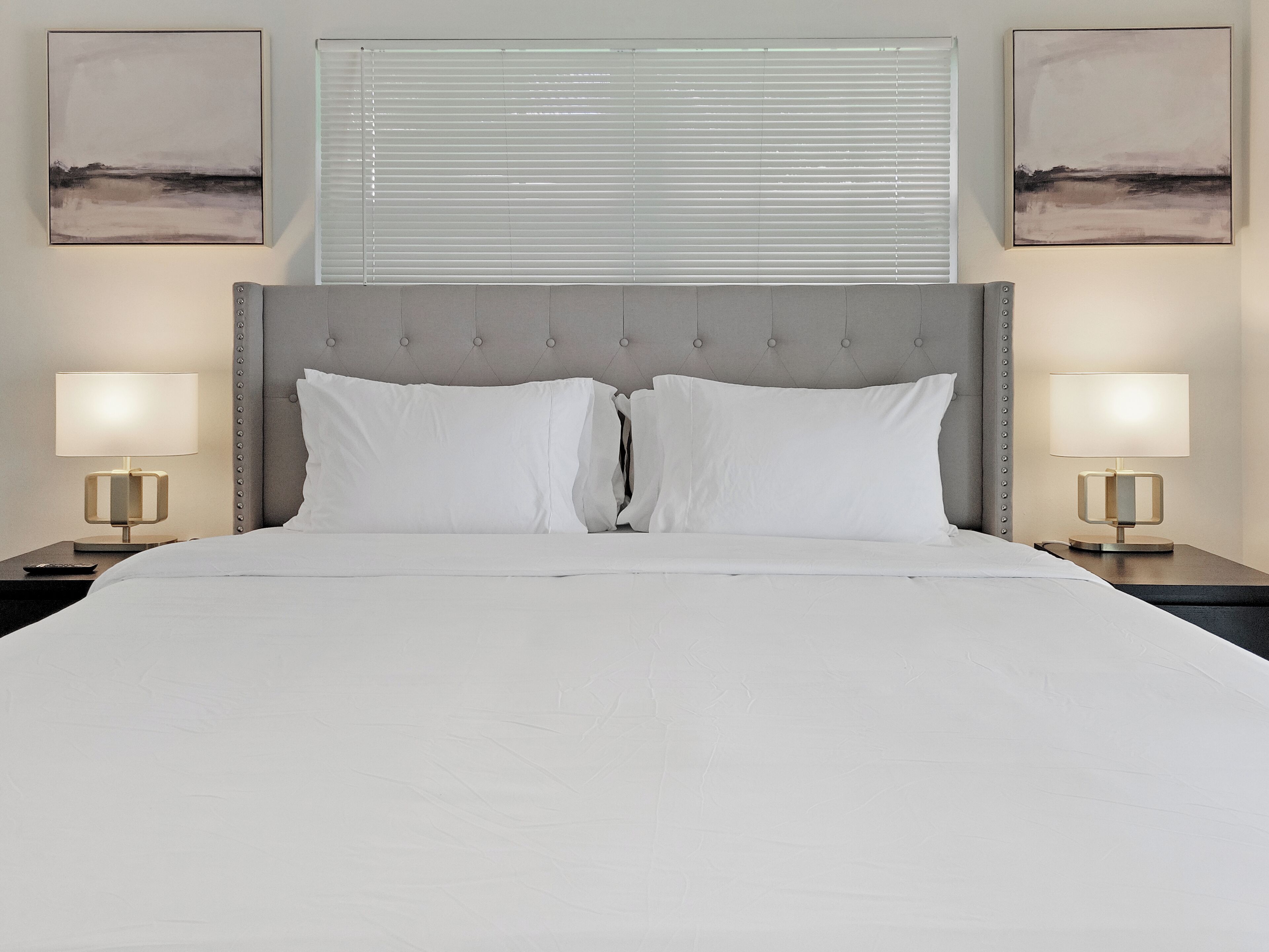 Un lit bien fait, avec du linge de lit blanc, une tête de lit rembourrée de couleur grise et deux lampes de chevet de chaque côté.