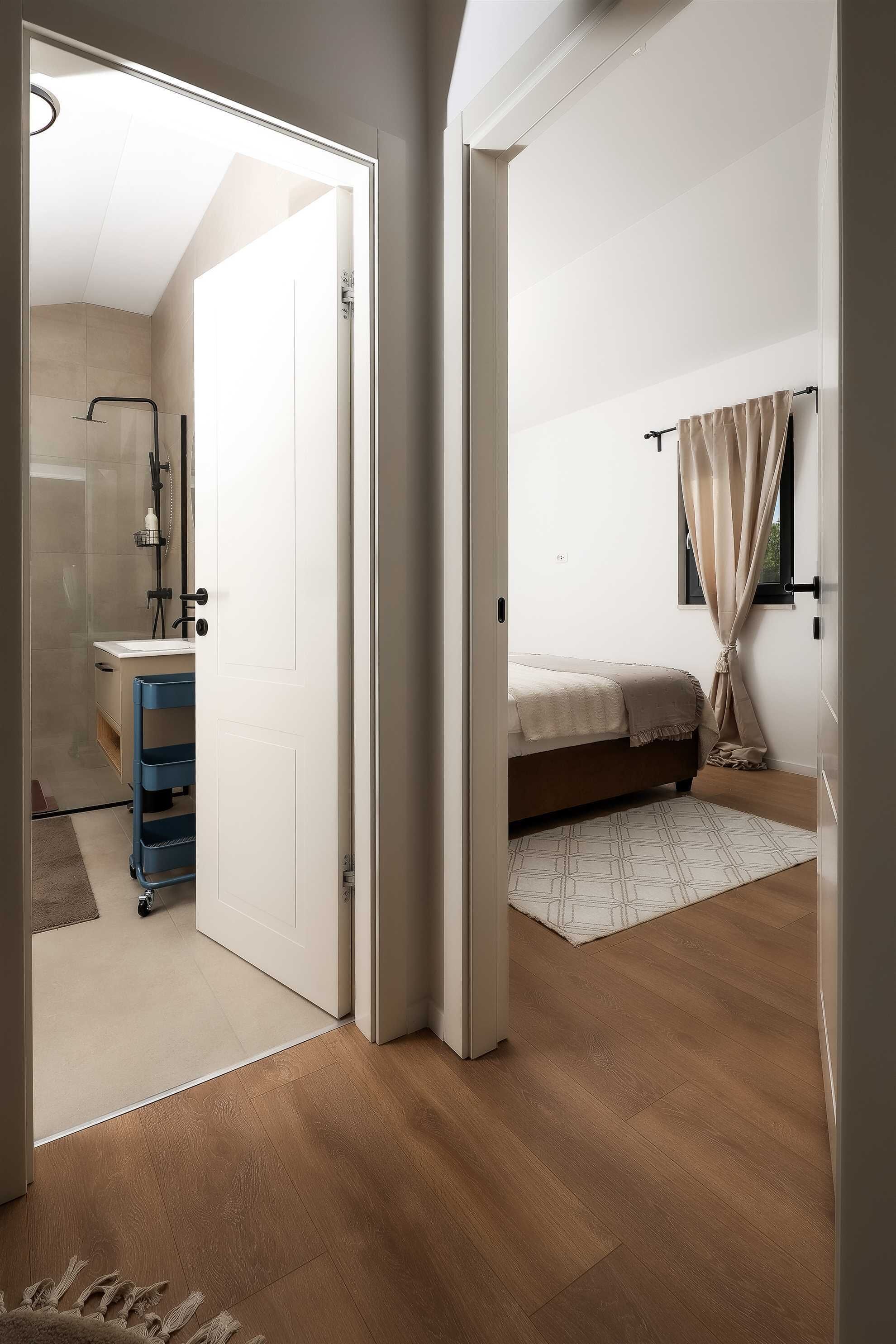 Un dormitorio con cama, ventana con cortinas, mesita de noche y puerta que da a un baño.
