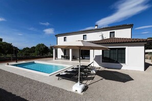 Una casa de dos pisos con piscina, mobiliario exterior y un patio cubierto.