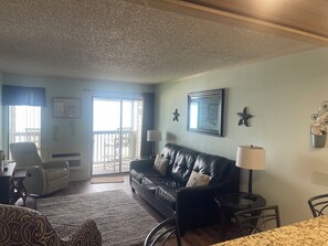Ein Wohnzimmer mit einer schwarzen Ledersofa, einem Esstisch und einem Stuhl. Eine Glastür führt auf einen Balkon.