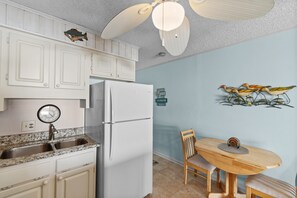 A kitchen with white cabinets, a white refrigerator, a sink, a clock, and a wall-mounted bird decoration. - Sun-kissed Dunes: 2-bedroom condo Oceanfront; pool sleeps 6 (Myrtle Beach)