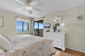 Ein Schlafzimmer mit Bett, Kommode, Deckenventilator, Fenster mit Jalousien und Blick ins Freie.