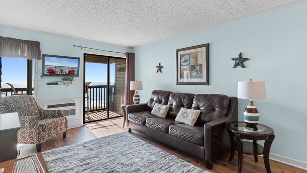 A living room with a leather couch, a patterned armchair, a glass coffee table, and a view of the ocean through a sliding door. - Sun-kissed Dunes: 2-bedroom condo Oceanfront; pool sleeps 6 (Myrtle Beach)