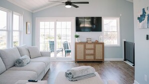 A living room with a sofa, a television, a ceiling fan, and a view through a glass door. - High Tides & Pink Skies-3BR, Pool, Views, Linens, Steps to Beach - North Topsail (North Topsail Beach)