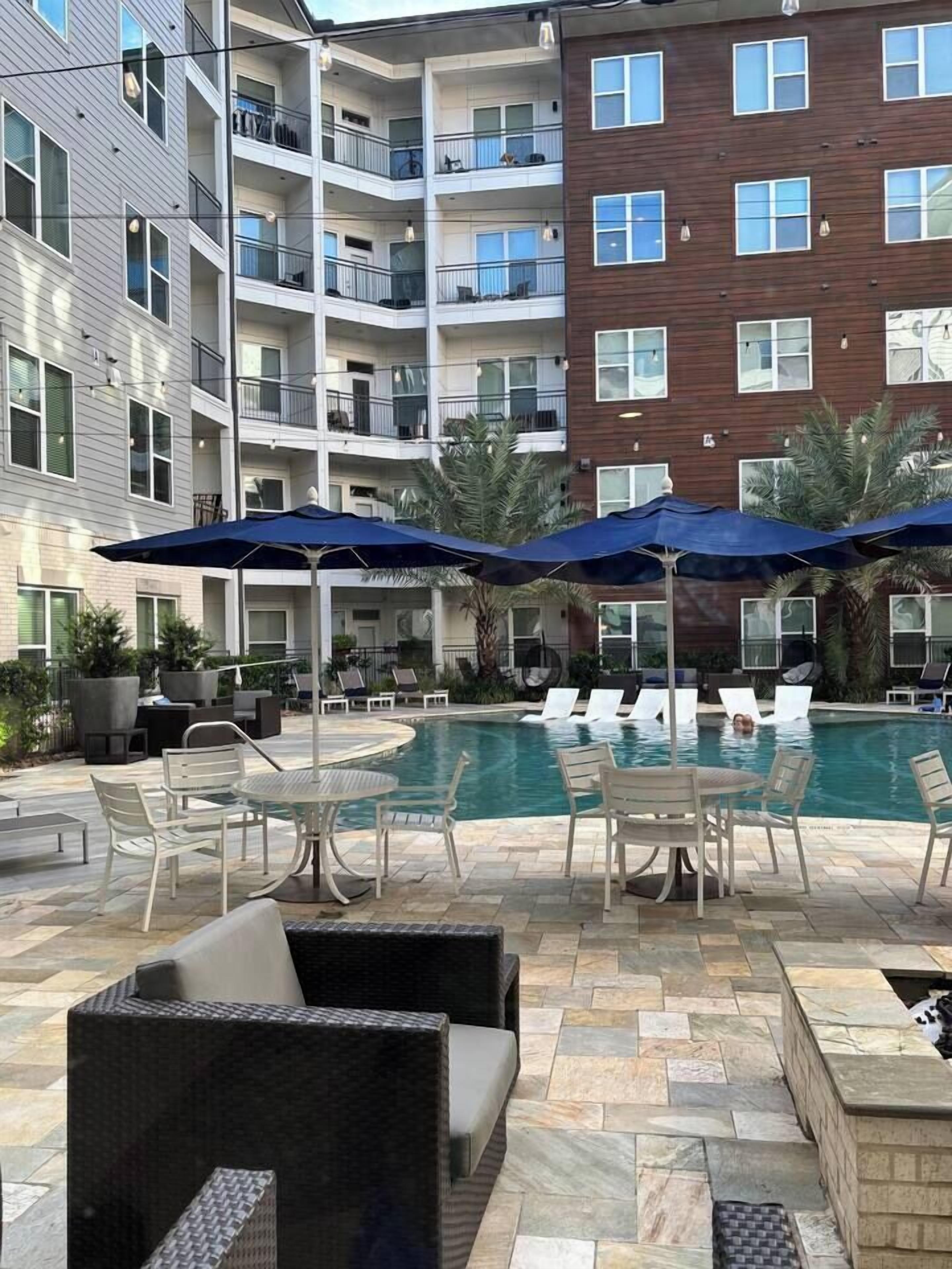 Un espace aménagé au bord de la piscine, avec des chaises longues et des parasols, entouré d’immeubles à plusieurs étages dotés de balcons.