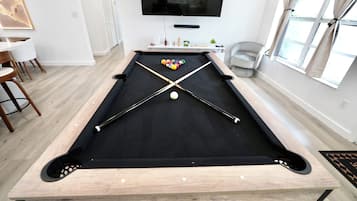 Una mesa de billar con taco y bolas, un televisor montado en la pared y una ventana con cortinas.