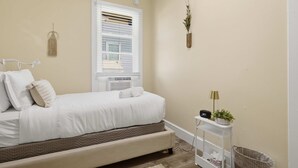 A bedroom with a bed, bedside table, lamp, radiator, window, wall decor, and a trash bin. - Charming Del Ray Home, Near Old Town & METRO-rail (Alexandria)