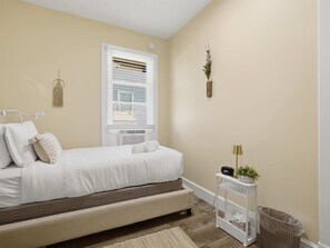 A bedroom with a bed, bedside table, lamp, radiator, window, wall decor, and a trash bin. - Charming Del Ray Home, Near Old Town & METRO-rail (Alexandria)