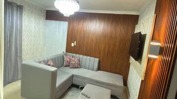 Una sala de estar con sofá modular, un pequeño reposapiés, un ventilador de techo y un televisor montado en la pared.