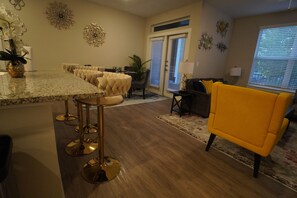Ein modernes Wohnzimmer mit einem gelben Sessel, eine Essecke mit einem langen Tisch und Stühlen sowie eine Küche mit einer Granit Arbeitsplatte.
