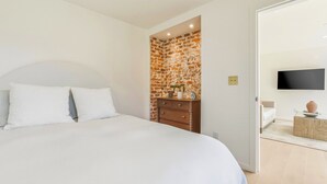 A bedroom with a large bed, a wooden dresser, a brick accent wall, and a flat-screen TV mounted on the wall. - Coastal Sanctuary: Newly Remodeled, Unrivaled Views, Sauna, and Steps to Ferry! (Sausalito)